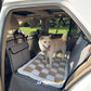 A light cream Shiba Inu dog stands on a checkered gray and beige dog car bed installed in the back seat of a white vehicle, with a grassy lawn visible through the rear window.